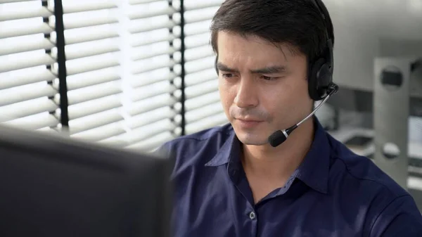 Young competent asian male call center agent working at his computer while simultaneously speaking with customers. Concept of an operator, customer service agent working in the office with headset.