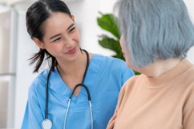 A caregiver rest her hands on the shoulders of a contented senior patient while she sitting on the sofa at home.