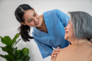 A caregiver rest her hands on the shoulders of a contented senior patient while she sitting on the sofa at home.