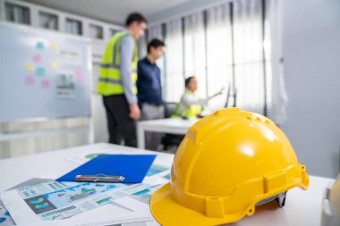 Concept of engineering equipments of protective helmet for competent engineer on piles of paper works. Blurred image of engineers and employer working together at the desk.