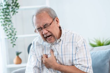 An agonizing senior man suffering from chest pain or heart attack alone in his living room. Serious health problem and feeling unwell concept for seniors.