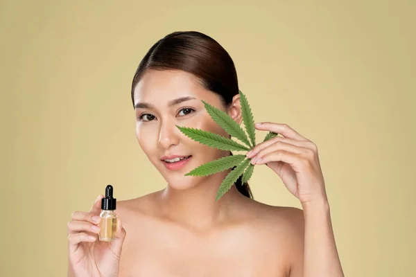 Portrait of young ardent woman with healthy fresh skin holding green hemp leaf and cbd oil. Combination of beauty and cannabis concept.