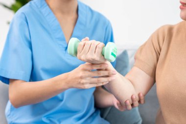 Caregiver helping contented senior woman exercise with dumbbell at home. Rehab for elder, senior medical care.