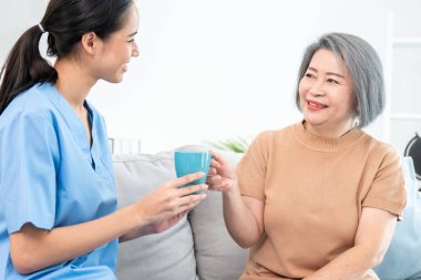 Female care taker serving her contented senior patient with a cup of coffee at home, smiling to each other. Medical care for pensioners, Home health care service.