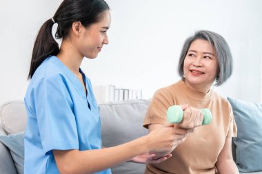 Caregiver helping contented senior woman exercise with dumbbell at home. Rehab for elder, senior medical care.