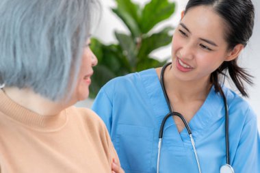 A caregiver rest her hands on the shoulders of a contented senior patient while she sitting on the sofa at home.