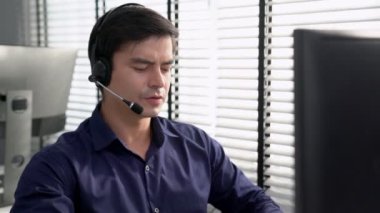 Young competent asian male call center agent working at his computer while simultaneously speaking with customers. Concept of an operator, customer service agent working in the office with headset.