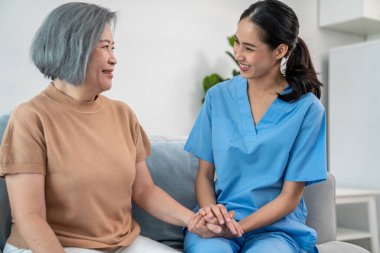 A caregiver rest her hands on the shoulders of a contented senior patient while she sitting on the sofa at home.