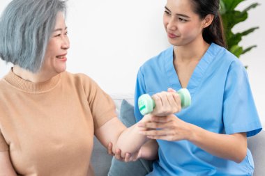 Caregiver helping contented senior woman exercise with dumbbell at home. Rehab for elder, senior medical care.