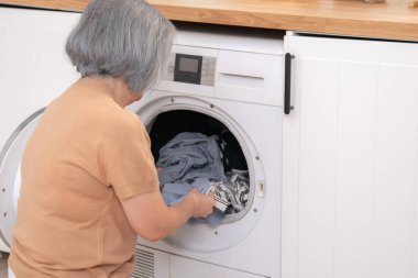 Contented senior housewife doing laundry in the laundry room with clothes inside the washing machine. Domestic life, drying machine, household chores.