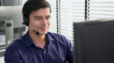 Young competent asian male call center agent working at his computer while simultaneously speaking with customers. Concept of an operator, customer service agent working in the office with headset.