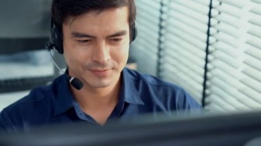 Young competent asian male call center agent working at his computer while simultaneously speaking with customers. Concept of an operator, customer service agent working in the office with headset.