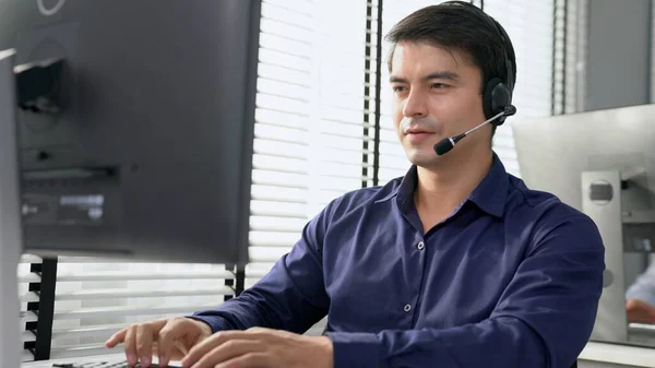 Young competent asian male call center agent working at his computer while simultaneously speaking with customers. Concept of an operator, customer service agent working in the office with headset.