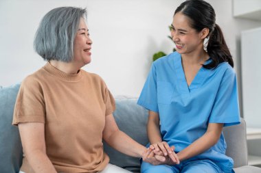 A caregiver rest her hands on the shoulders of a contented senior patient while she sitting on the sofa at home.
