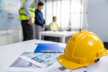 Concept of engineering equipments of protective helmet for competent engineer on piles of paper works. Blurred image of engineers and employer working together at the desk.