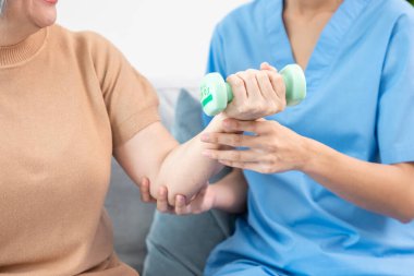 Caregiver helping contented senior woman exercise with dumbbell at home. Rehab for elder, senior medical care.