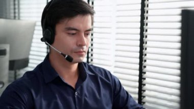 Young competent asian male call center agent working at his computer while simultaneously speaking with customers. Concept of an operator, customer service agent working in the office with headset.