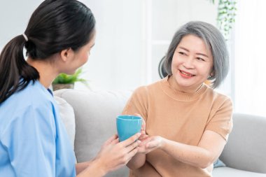Female care taker serving her contented senior patient with a cup of coffee at home, smiling to each other. Medical care for pensioners, Home health care service.