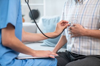 Caring young female doctor examining her contented senior patient with stethoscope in living room. Medical service for elderly, elderly sickness, declining health.