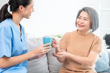 Female care taker serving her contented senior patient with a cup of coffee at home, smiling to each other. Medical care for pensioners, Home health care service.