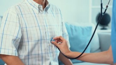 Caring young female doctor examining her contented senior patient with stethoscope in living room. Medical service for elderly, elderly sickness, declining health.