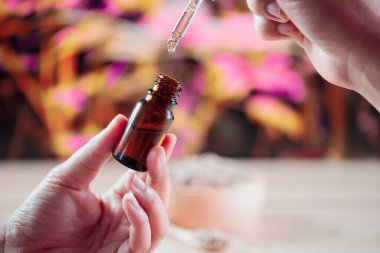 Eller, arka planda kenevir yaprağı olan bir şişe CBD yağı ve damlalık kapağını tutuyor. Tıbbi amaçlar için yasallaştırılmış CBD ürünü.