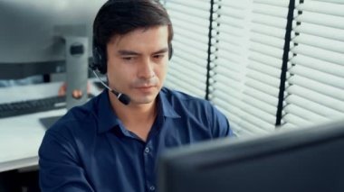 Young competent asian male call center agent working at his computer while simultaneously speaking with customers. Concept of an operator, customer service agent working in the office with headset.