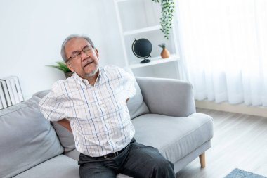 An agonizing senior man in need of assistance while sitting on his sofa at home, suffering from back pain. Senior care, nursing home for pensioners, deteriorating health of old age.