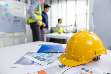 Concept of engineering equipments of protective helmet for competent engineer on piles of paper works. Blurred image of engineers and employer working together at the desk.