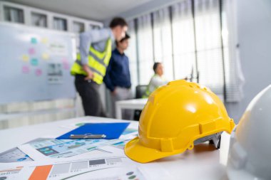 Concept of engineering equipments of protective helmet for competent engineer on piles of paper works. Blurred image of engineers and employer working together at the desk.