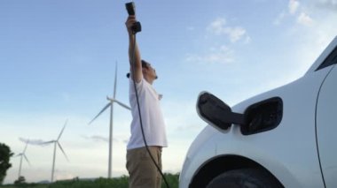 Elektrikli arabasıyla ilerici bir adam, EV arabası gelecekteki sürdürülebilir enerji kavramı olarak rüzgar türbiniyle yeşil alandaki enerji şarj etme istasyonundan enerji yüklüyor. Enerji üreteci olan elektrikli bir araç..