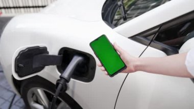 Taklit uzay için şarj istasyonuna bağlı elektrikli aracın yeşil ekran enerji durumunu gösteren bir model telefon. Temiz çevre için ilerici bir kavram. EV, yeşil yenilenebilir enerjiden güç alıyor.