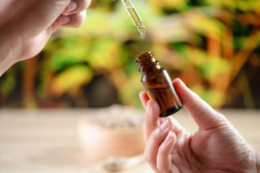 Eller, arka planda kenevir yaprağı olan bir şişe CBD yağı ve damlalık kapağını tutuyor. Tıbbi amaçlar için yasallaştırılmış CBD ürünü.