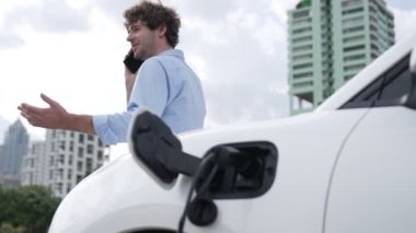 Takım elbiseli, hırslı bir işadamı elektrik kablosu ve yenilenebilir enerji enerjili bir elektrikli araçla şarj istasyonunda dururken elektrikli aracına yaslanıyor..