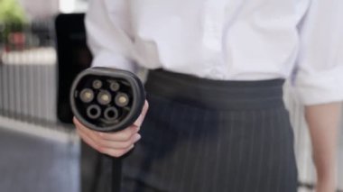 İlerici iş kadını şehir merkezinde dolaşmadan önce şarj cihazından elektrikli arabasına şarj prizi takıyor. Çevre dostu şarj edilebilir araba, sürdürülebilir ve temiz enerjiyle çalışıyor..