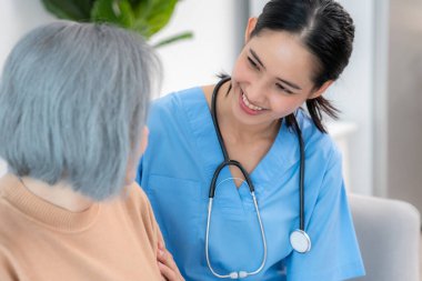 A caregiver rest her hands on the shoulders of a contented senior patient while she sitting on the sofa at home.