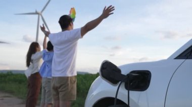 İlerici mutlu aile kavramı elektrikli araçla rüzgar çiftliğinde eğleniyor. Rüzgar türbini jeneratöründen gelen temiz yenilenebilir enerji ile şarj edilen elektrikli araç..