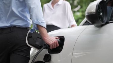 İlerici işadamı ve iş kadını, şehir merkezinde dolaşmadan önce şarj cihazını şarj istasyonundan elektrikli arabaya takıyor. Çevre dostu şarj edilebilir araba temiz enerjiyle çalışıyor..