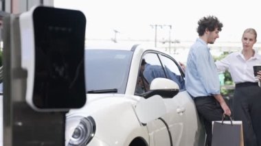 İlerici iş adamı ve iş kadını şehir merkezinde dolaşmadan önce şarj istasyonuna bağlı elektrikli arabaya yaslanıyor. Çevre dostu şarj edilebilir araba temiz enerjiyle çalışıyor..